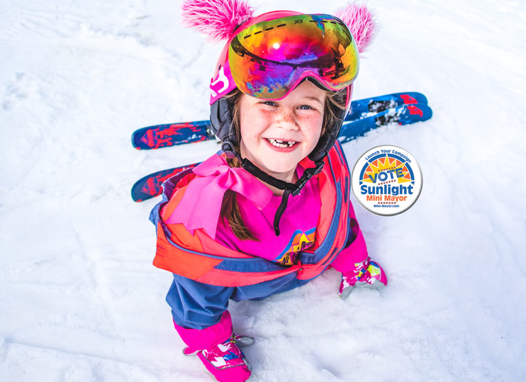 Girl with cute helmet on that has pink fluffy ears sits on the snow in her pink ski suit and smiles to the camera with her front teeth missing