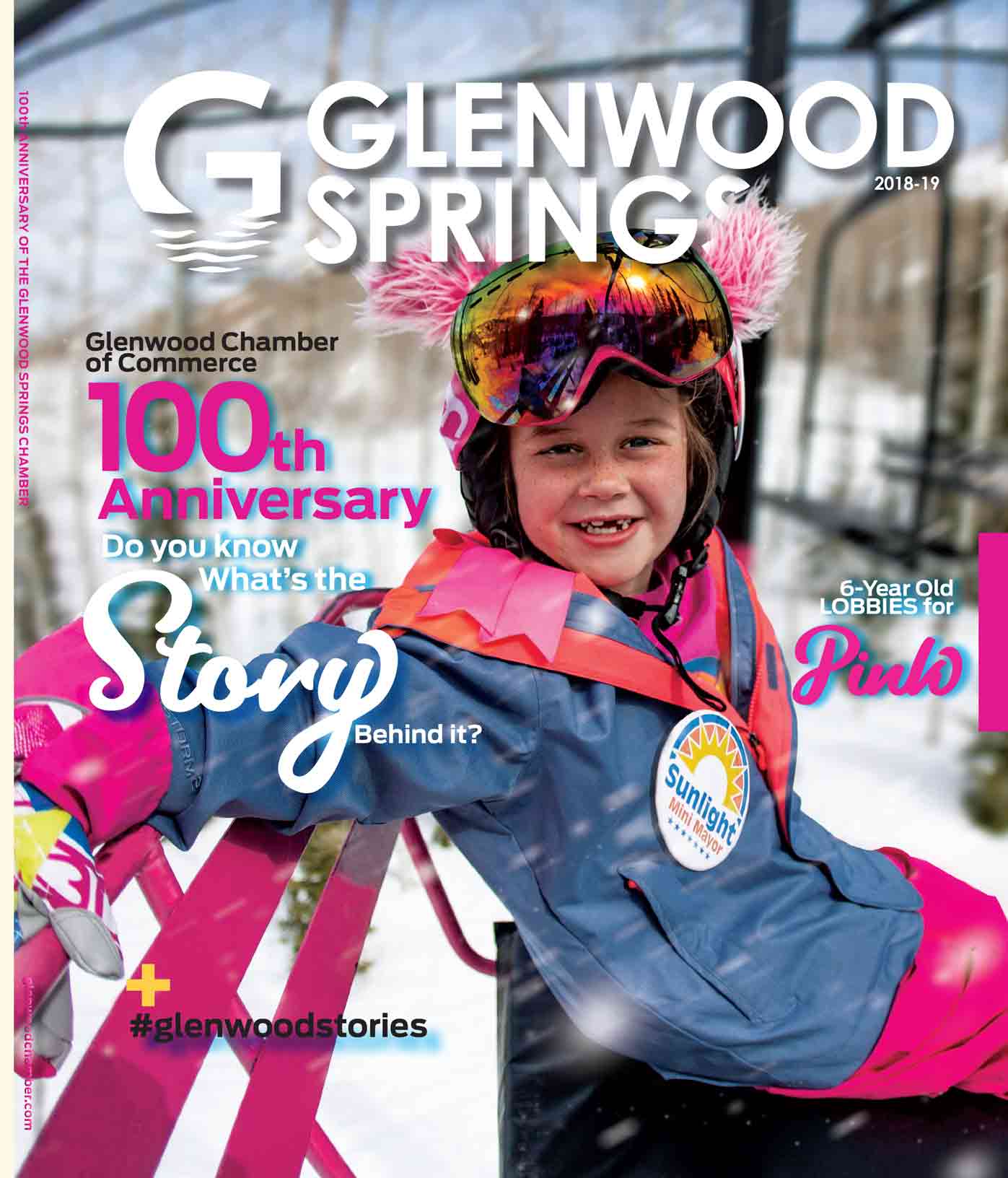 Cover of Glenwood springs magazine, a young girl is sitting on a pink chairlift with a blue jacket and pink pants and gloves, she is smiling towards the camera with missing teeth and a ski helmet on that has pink fluffy ears on it and pink googles