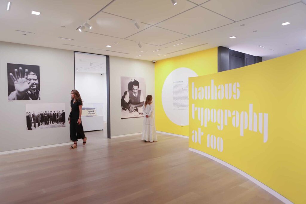 a view of a gallery with a curved wall that reads "Bayhaus typography at 100" two woman walk and observe the wall of art displays