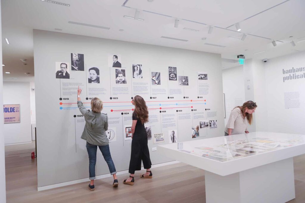 In a gallery room with a timeline of images and text three gallery observers glance and point at the timeline discussing the storyline