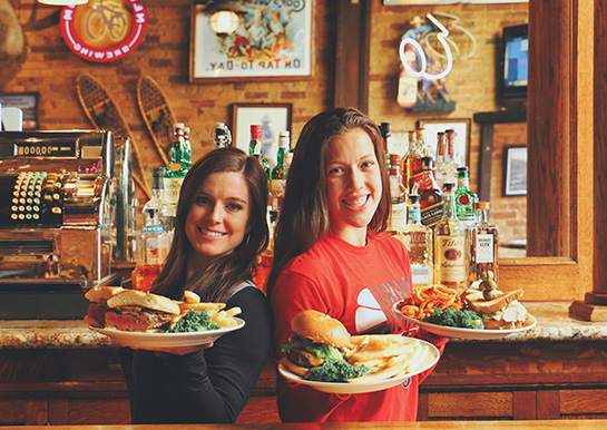 Two young beautiful woman stand behind a bar back to back each smiling and holding platters of food with a western bar in the background